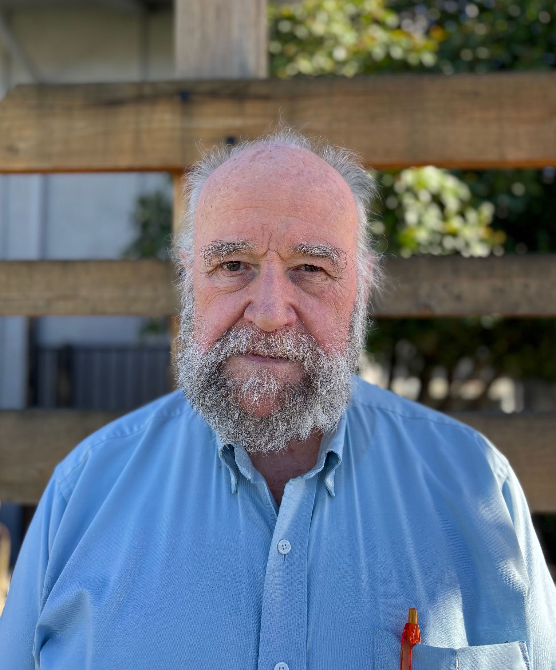 A man with a beard and a blue shirt is standing in front of a wooden fence.