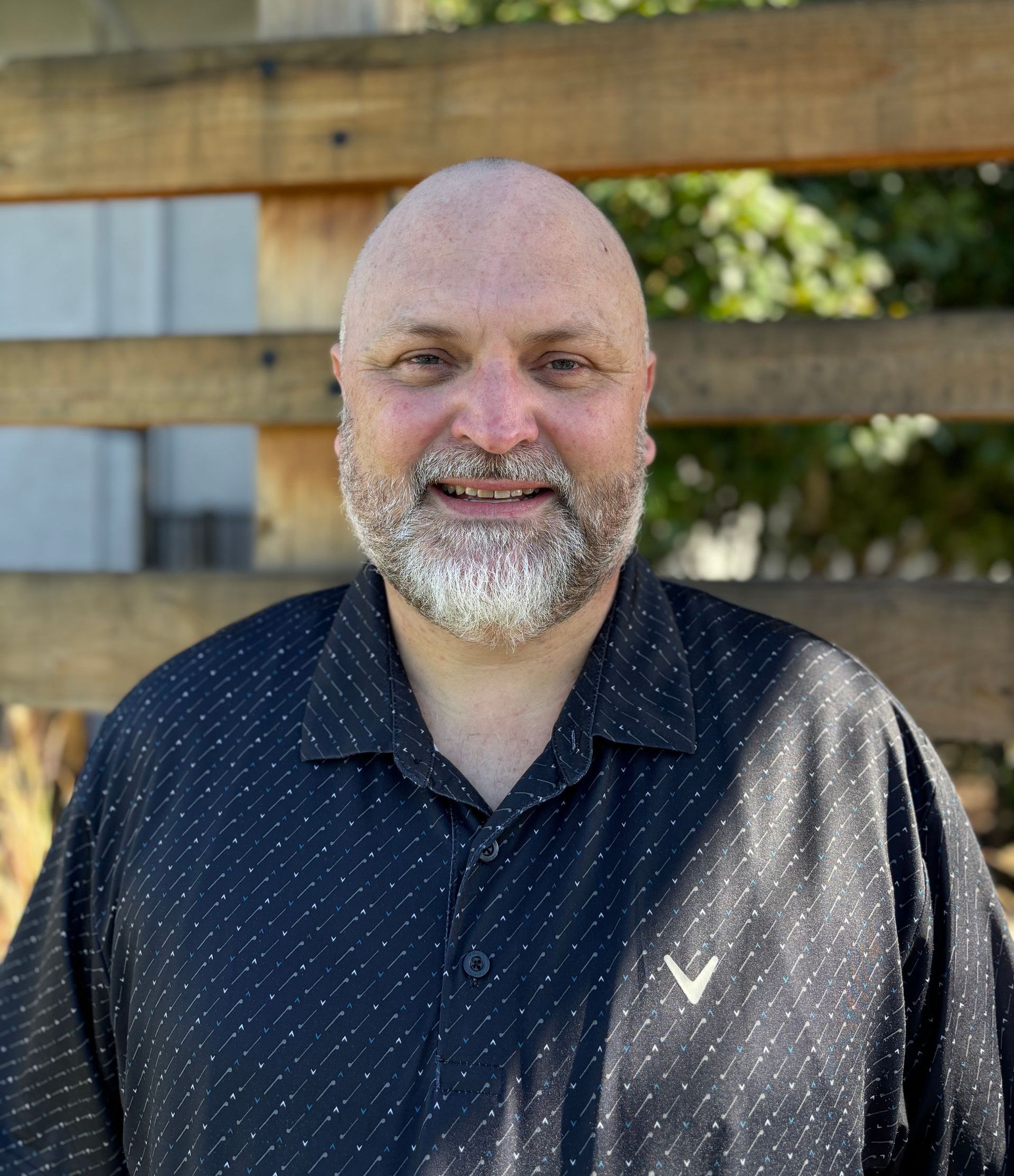 A bald man with a beard wearing a black shirt is standing in front of a wooden fence.