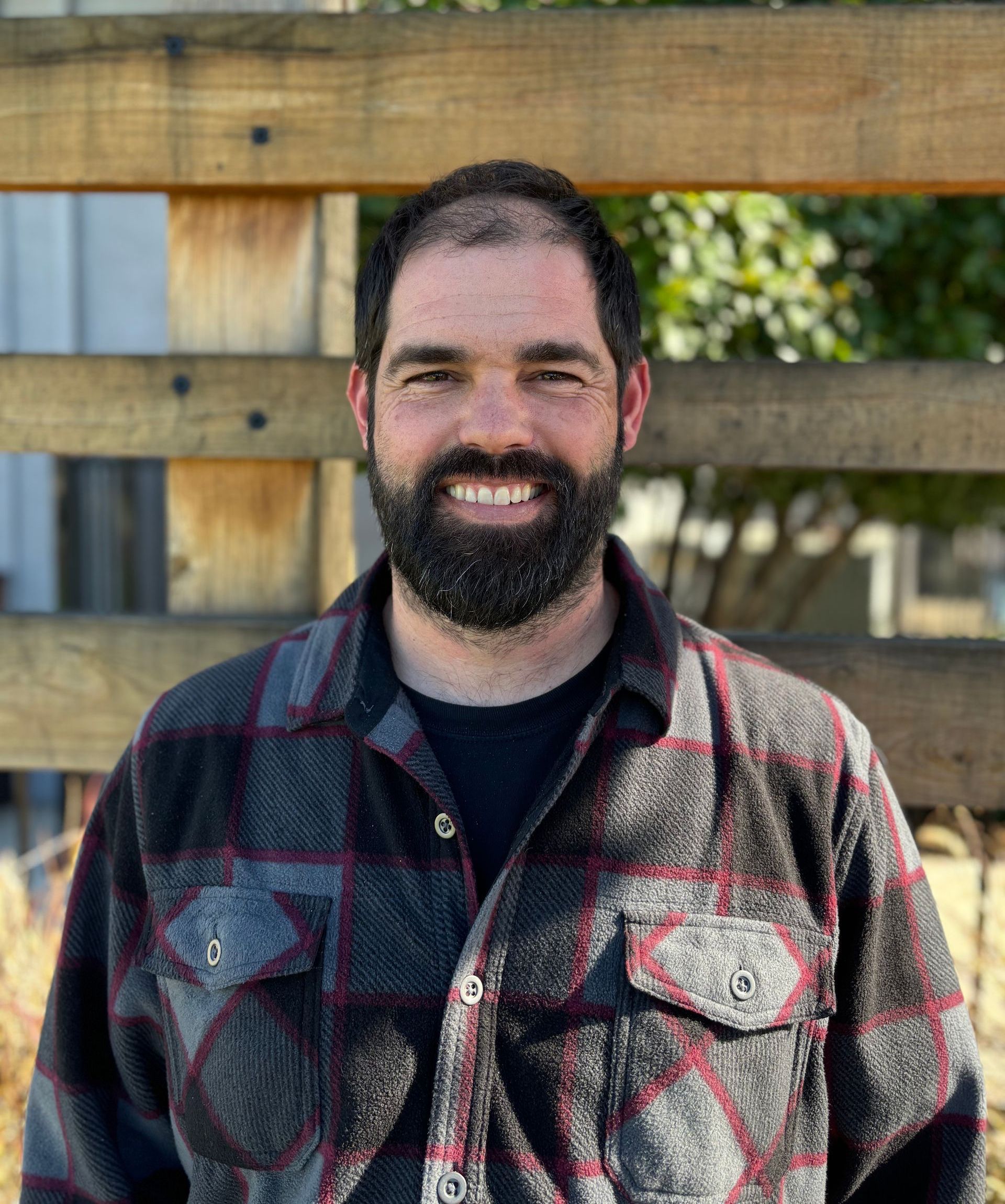 A man with a beard and a plaid shirt is smiling in front of a wooden fence.