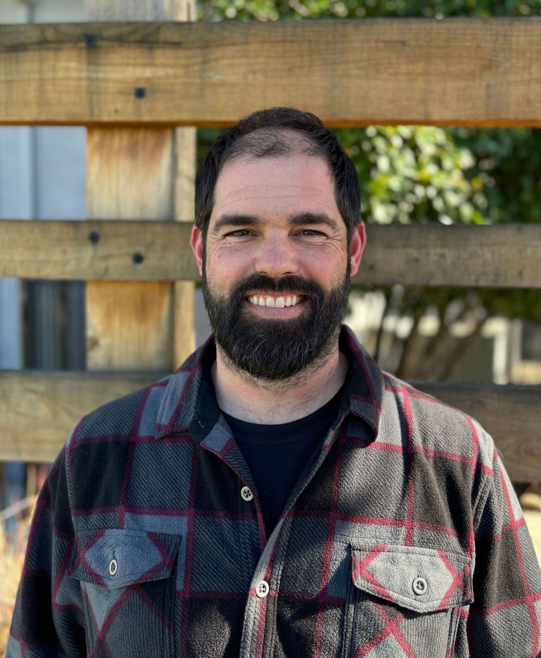 A man with a beard and a plaid shirt is smiling in front of a wooden fence.