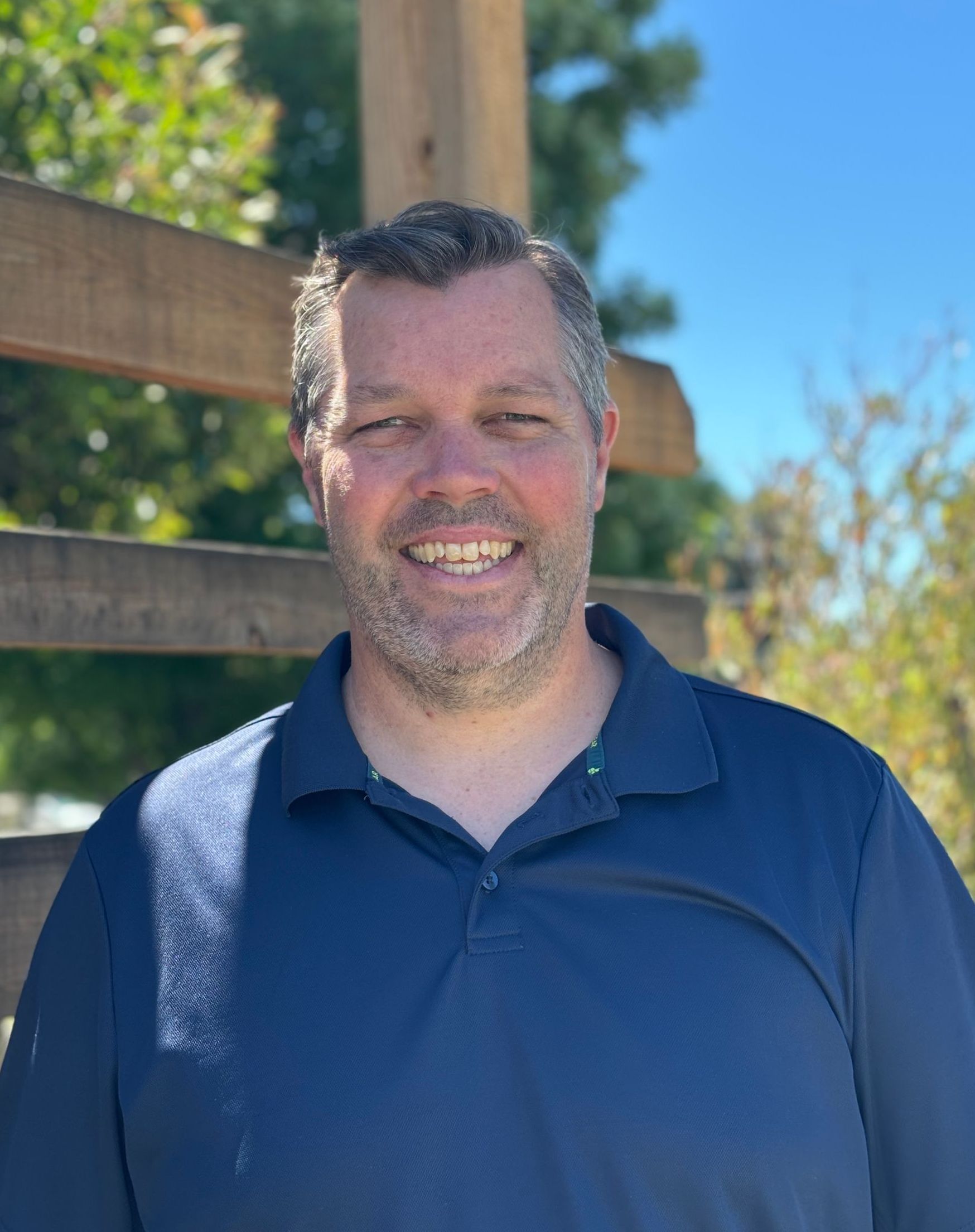 A man with a beard and a blue shirt is smiling in front of a wooden fence.