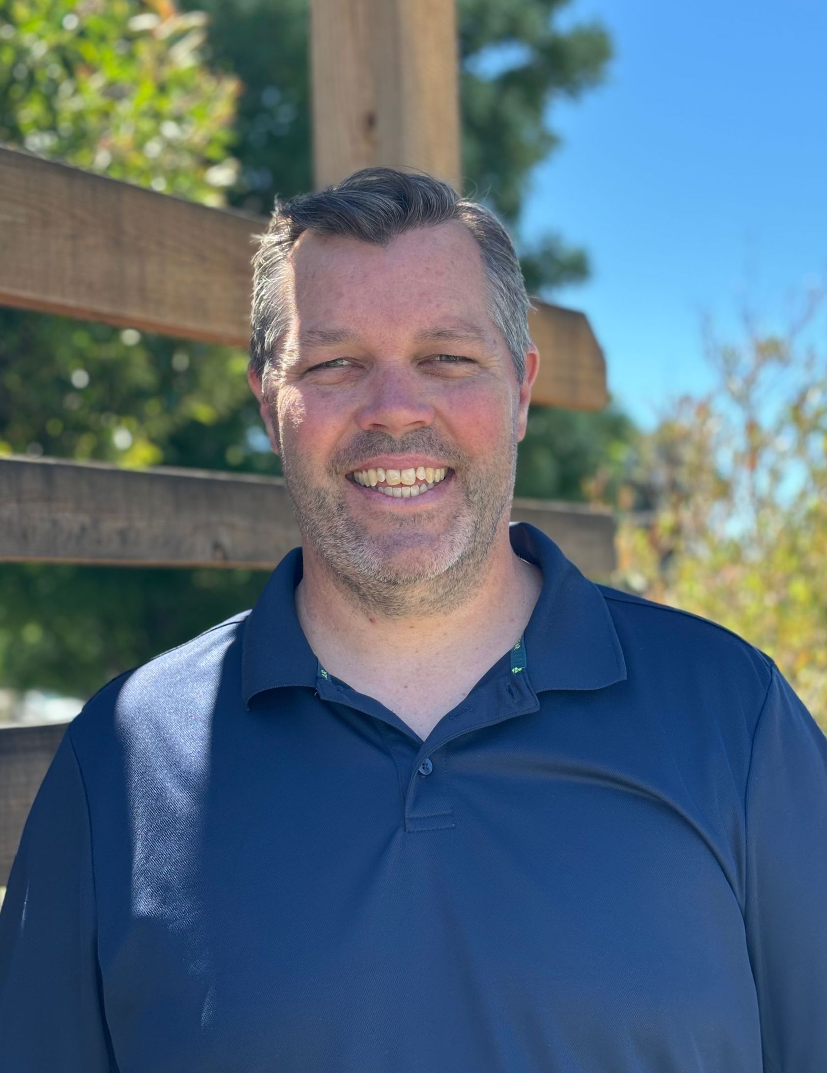 A man in a blue shirt is smiling in front of a wooden fence.