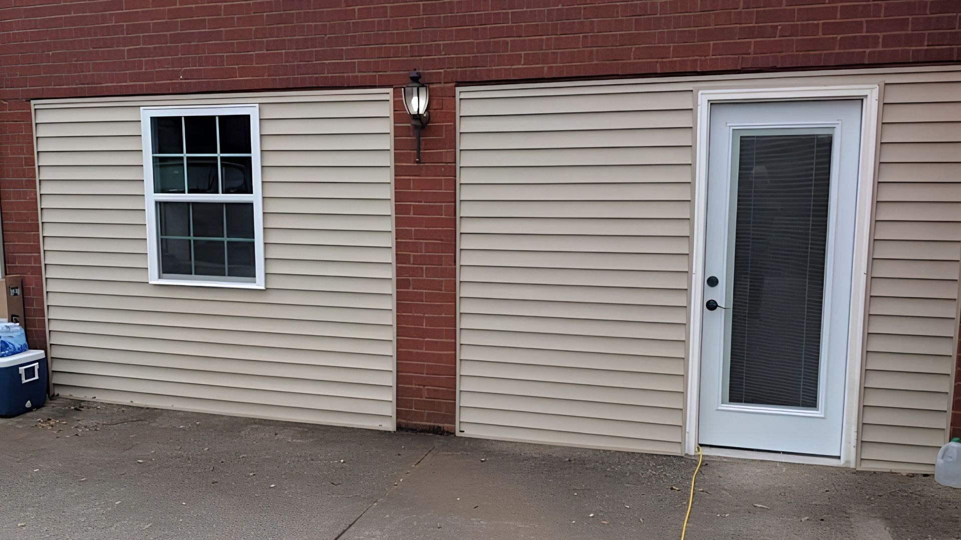 Beige siding on a brick building with a window, door, and outdoor light over a concrete walkway.