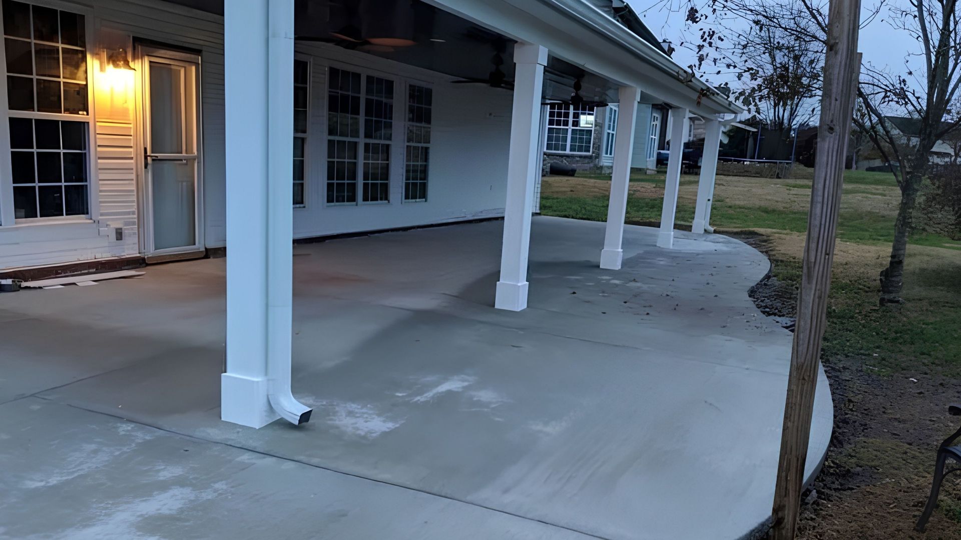 Covered concrete porch beside a white house at dusk, with lit entryway and columns