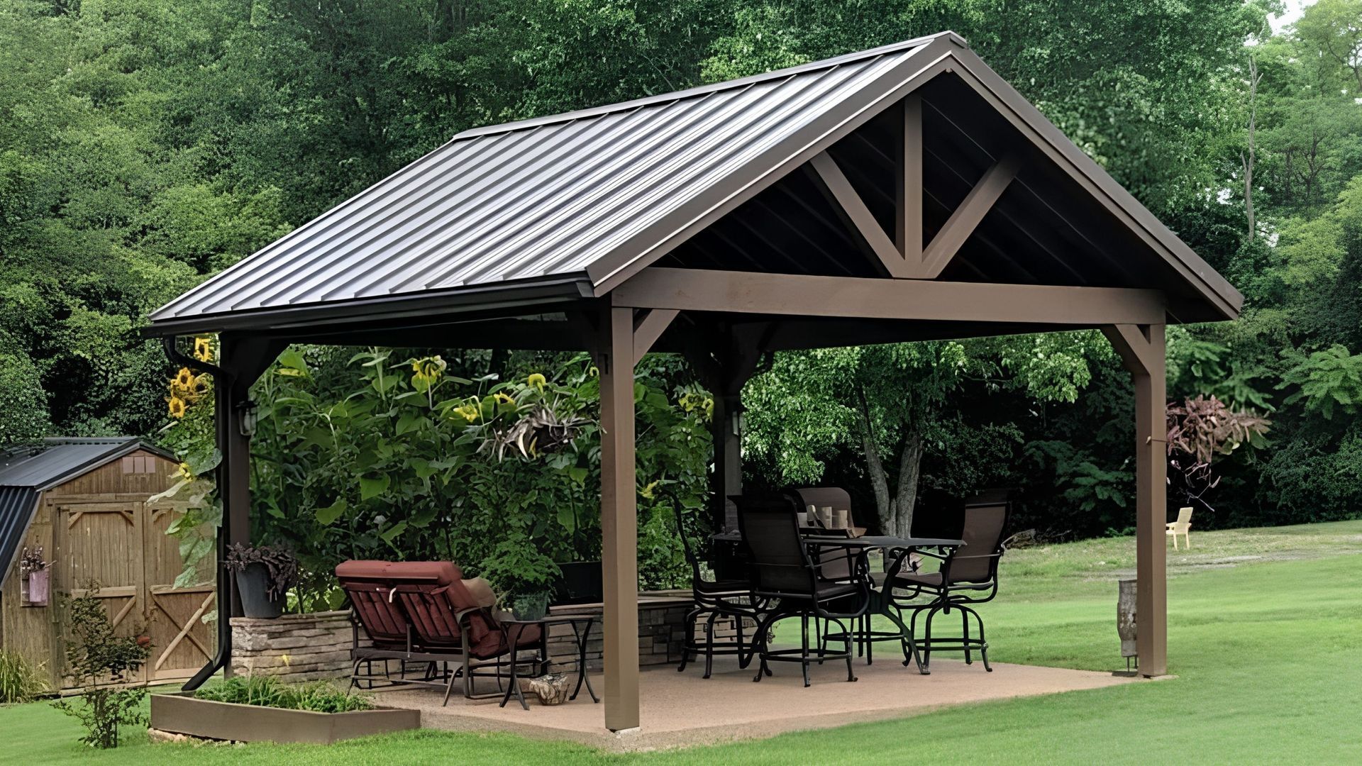 Open-sided backyard pavilion with metal roof, patio furniture, and green lawn surrounded by trees