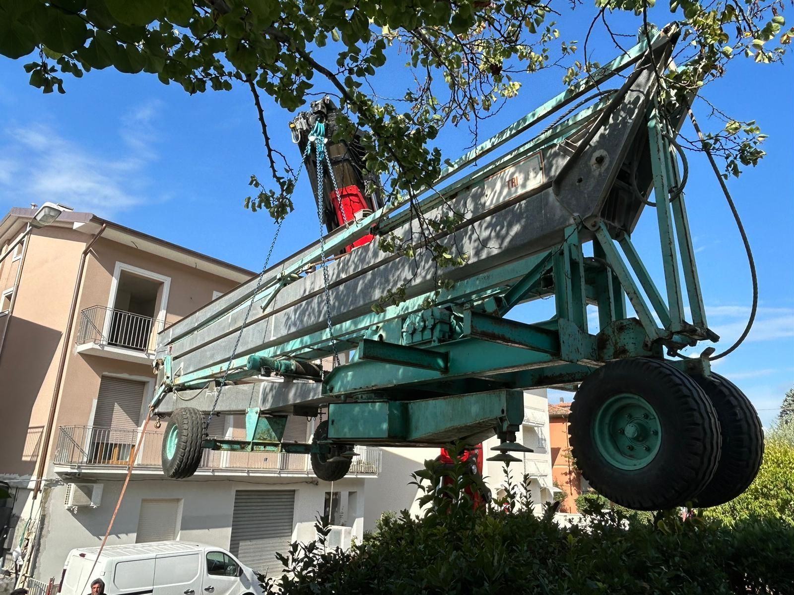 Una grande gru verde solleva una barca bianca sopra una casa in un quartiere soleggiato