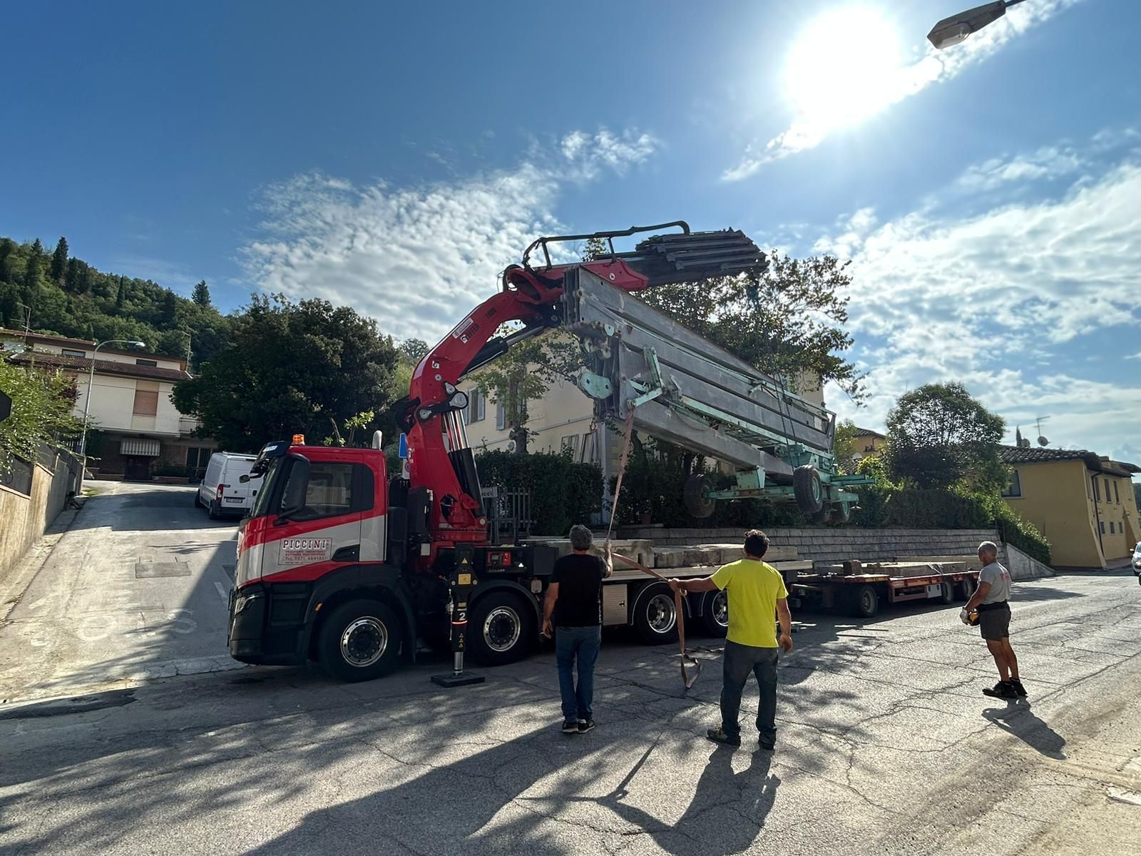 Un camion gru rosso solleva una grande struttura metallica su una strada soleggiata, con due operai nelle vicinanze.