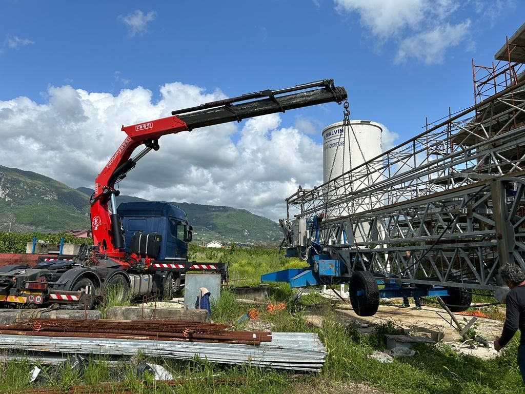 Una gru rossa solleva una travatura reticolare in acciaio in un sito industriale, con operai nelle vicinanze e un silo sullo sfondo.