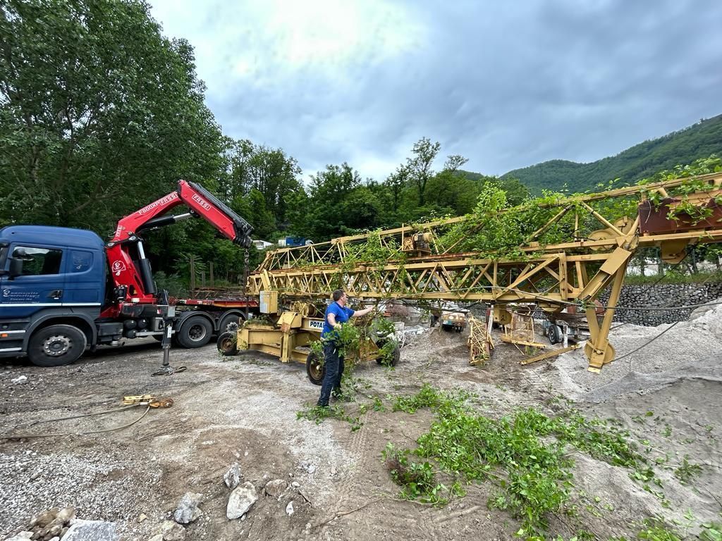 Un operaio, accanto a una gru montata su un camion, solleva legname in un cantiere roccioso all'aperto, tra alberi e colline.