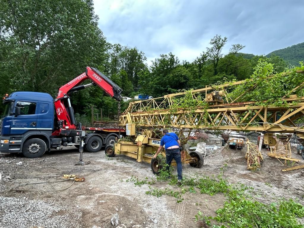 Un camion con gru rosso scarica una lunga travatura metallica accanto a un cantiere fangoso in una zona boschiva.