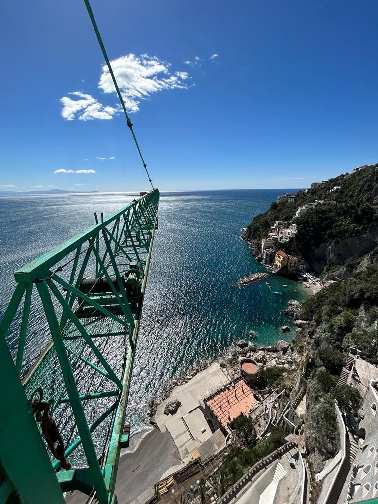 Vista dalla funivia su una baia turchese, scogliere rocciose e edifici sul lungomare sotto un cielo azzurro brillante.