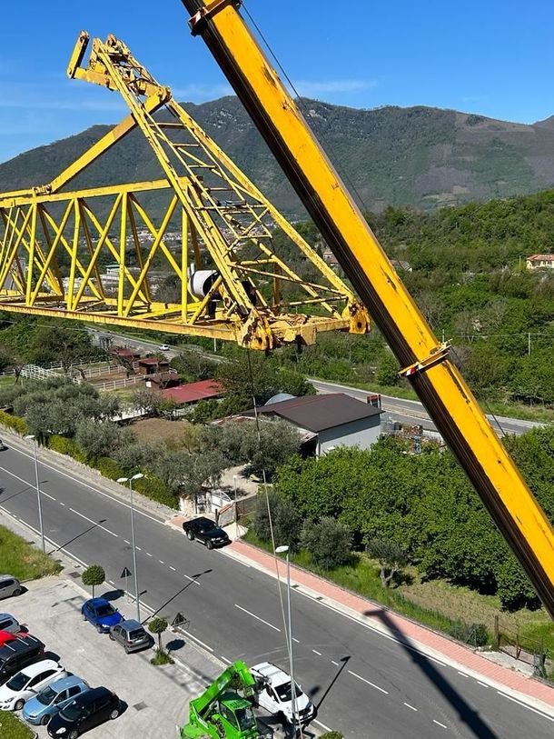 Una gru gialla solleva un grande segmento di ponte sopra una strada, tra alberi e montagne.