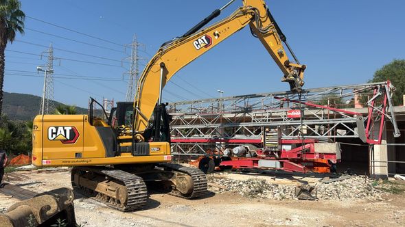 Un escavatore CAT giallo solleva una struttura metallica rossa in un cantiere all'aperto sotto un cielo azzurro.