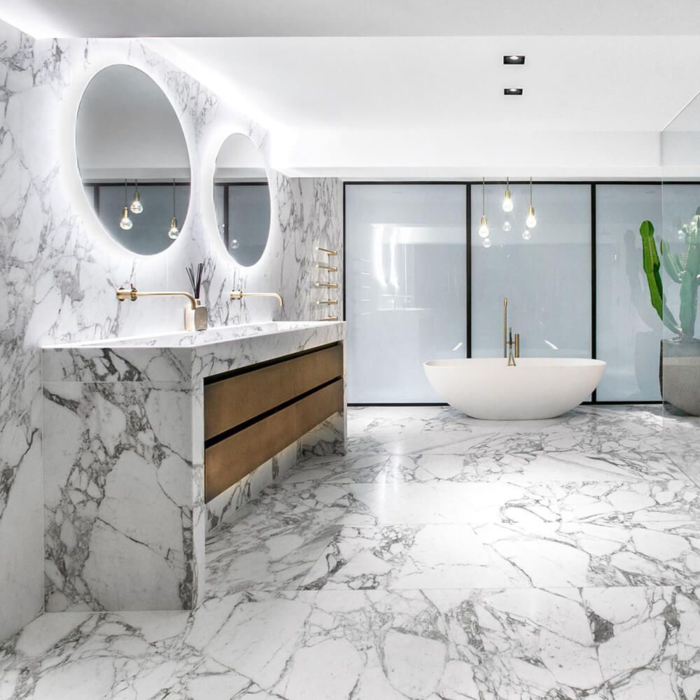 Modern white marble bathroom with oval mirrors, freestanding tub, and glass shower enclosure