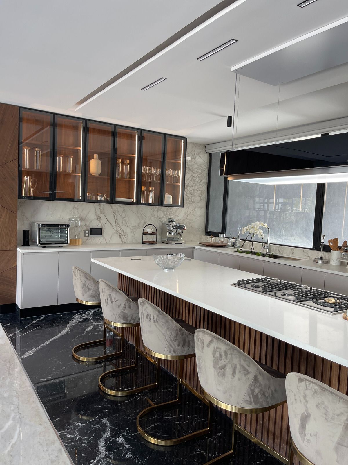 Modern kitchen with white island, marble counter, brass stools, dark floor, and glass-front cabinets