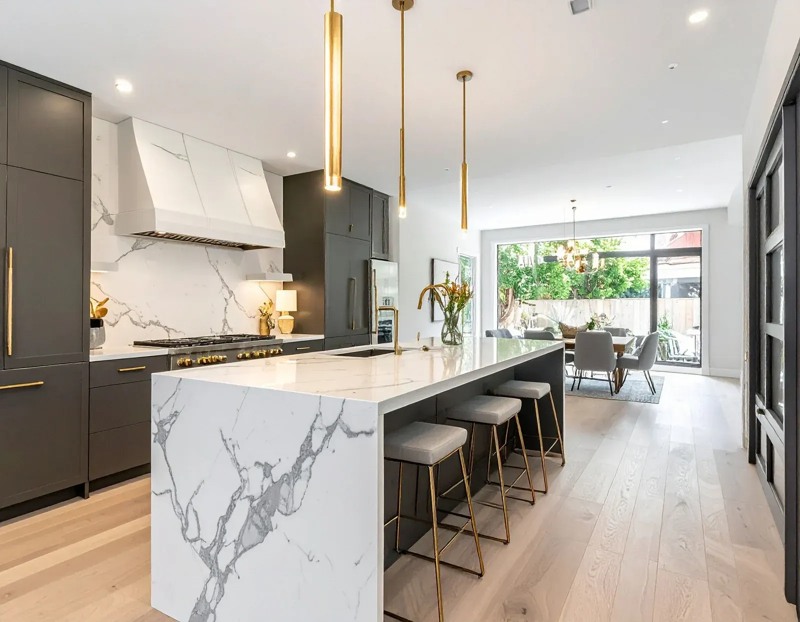 Modern white kitchen with marble island, black stools, gold pendant lights, and open dining area.