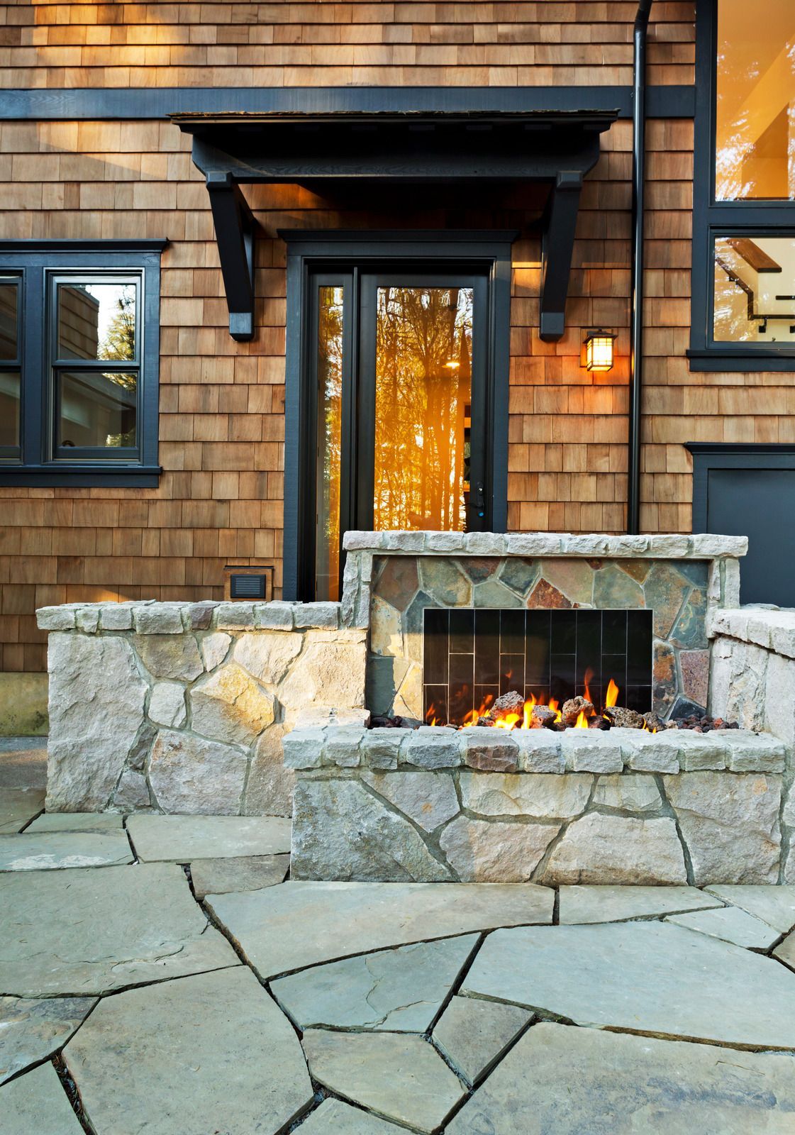 Stone fireplace with lit flames on a shingled house patio at dusk