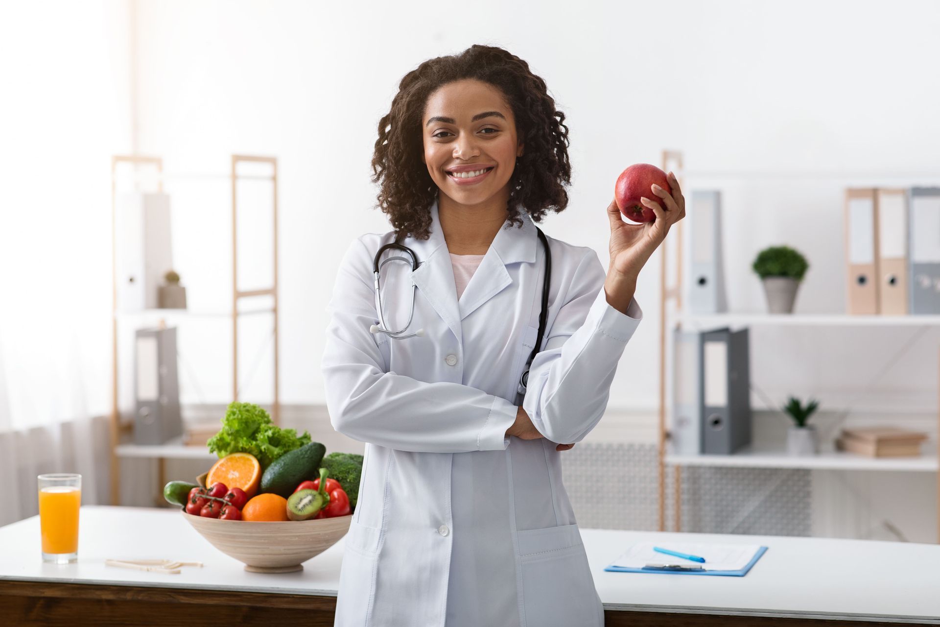 Uma médica está segurando uma maçã na mão em frente a uma tigela de frutas e vegetais.
