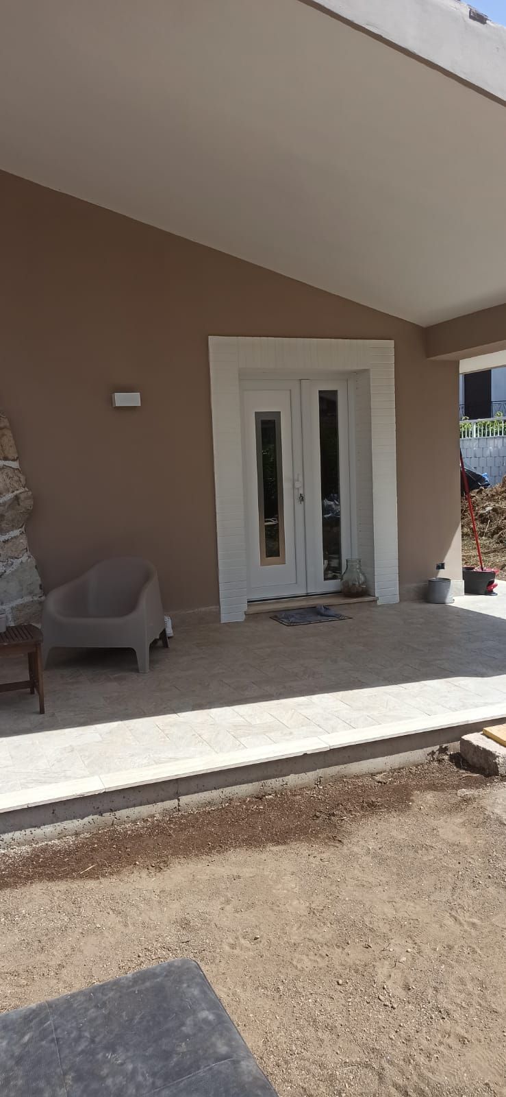 Vista esterna dell'ingresso di una casa con doppie porte e veranda coperta. L'edificio è dipinto in toni neutri.