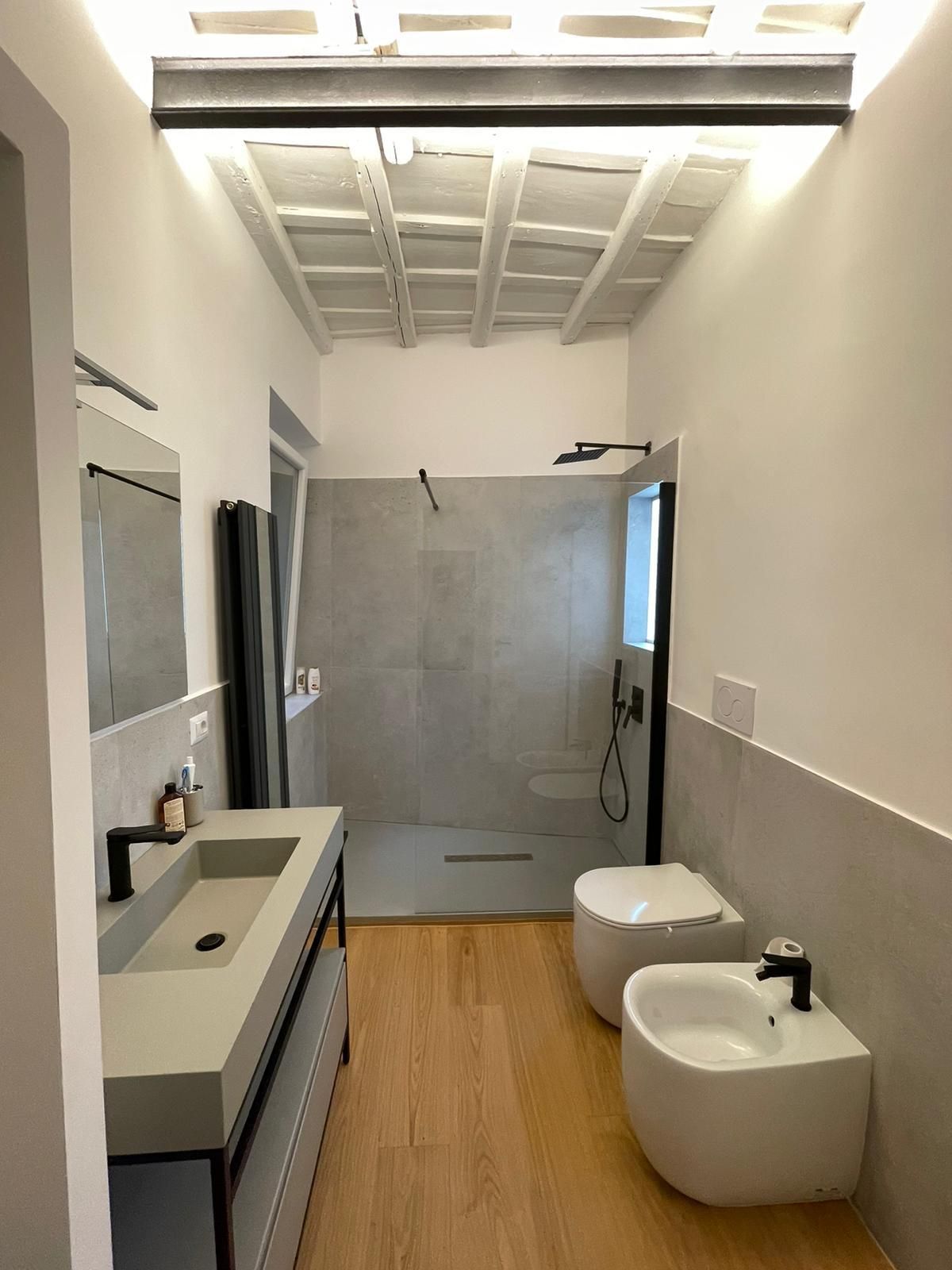 Bagno moderno con lavabo in cemento, cabina doccia, WC e bidet. Pavimento in legno, pareti bianche e travi a vista.