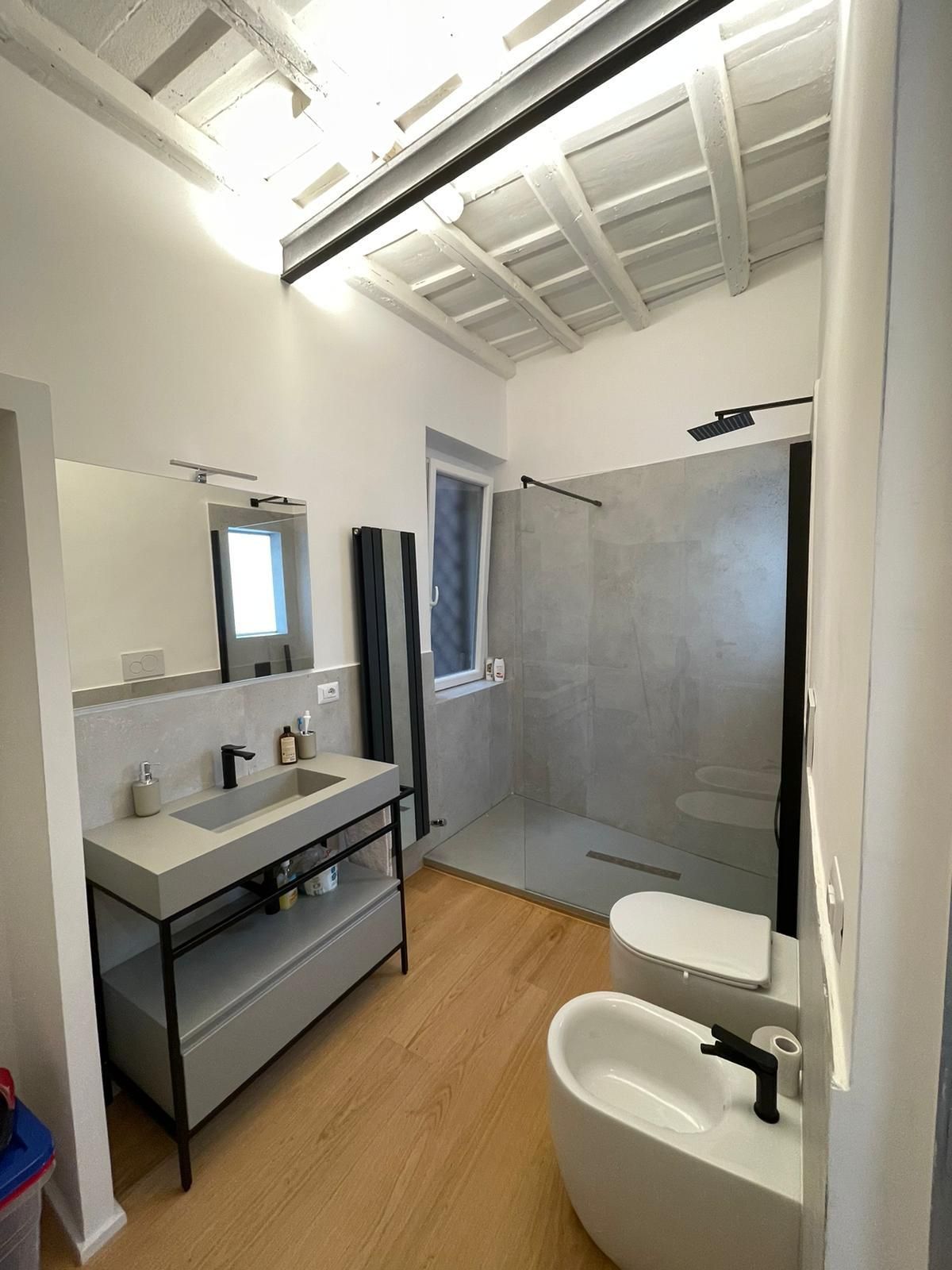 Bagno moderno con mobile lavabo in cemento, doccia, WC e bidet. Pavimento in legno, soffitto bianco e finestra.