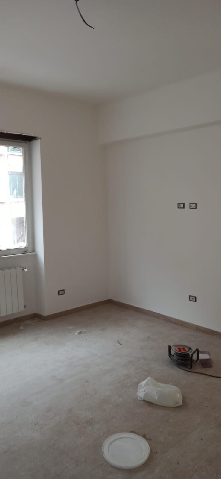 Una stanza vuota e incompiuta, con pareti bianche, una finestra e pavimento a vista. Materiali da costruzione sul pavimento.