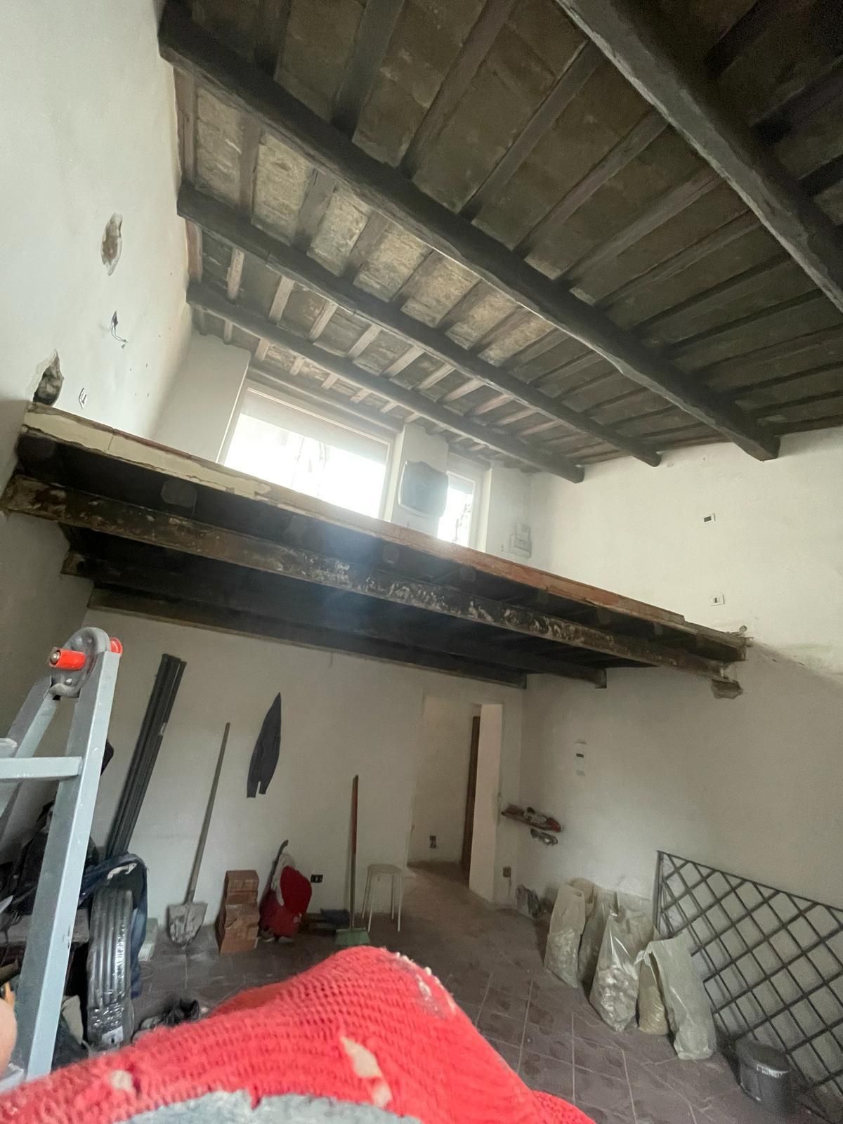 Interno di una stanza con travi in legno a vista, una scala e attrezzi. Pareti bianche e luce naturale.