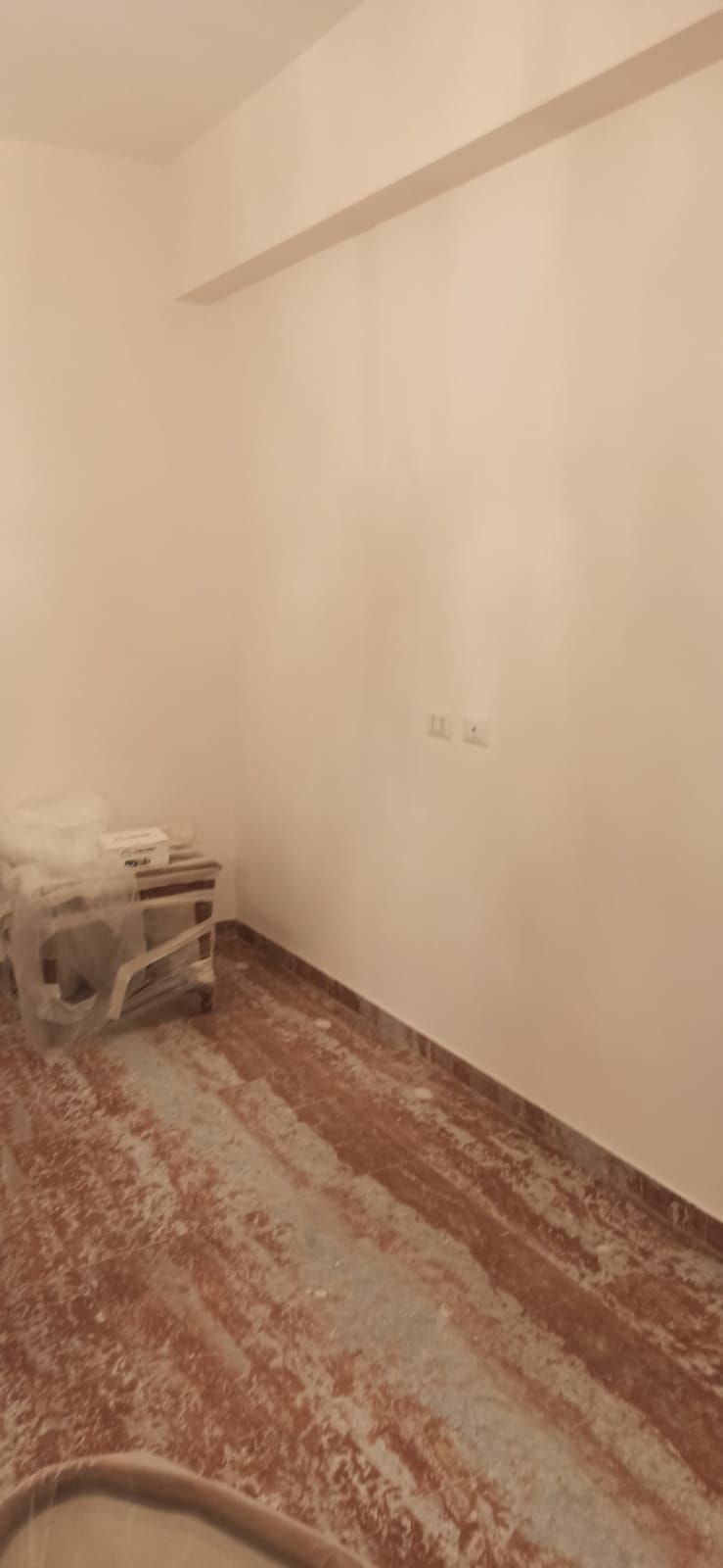 Una stanza vuota con pareti bianche e un pavimento rosso-marrone ricoperto di polvere. Un tavolino è appoggiato sulla sinistra.