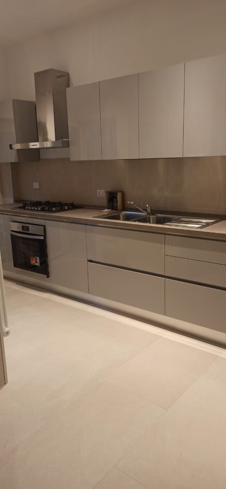 Cucina moderna con mobili grigio chiaro, cappa in acciaio inox e pavimento piastrellato chiaro.