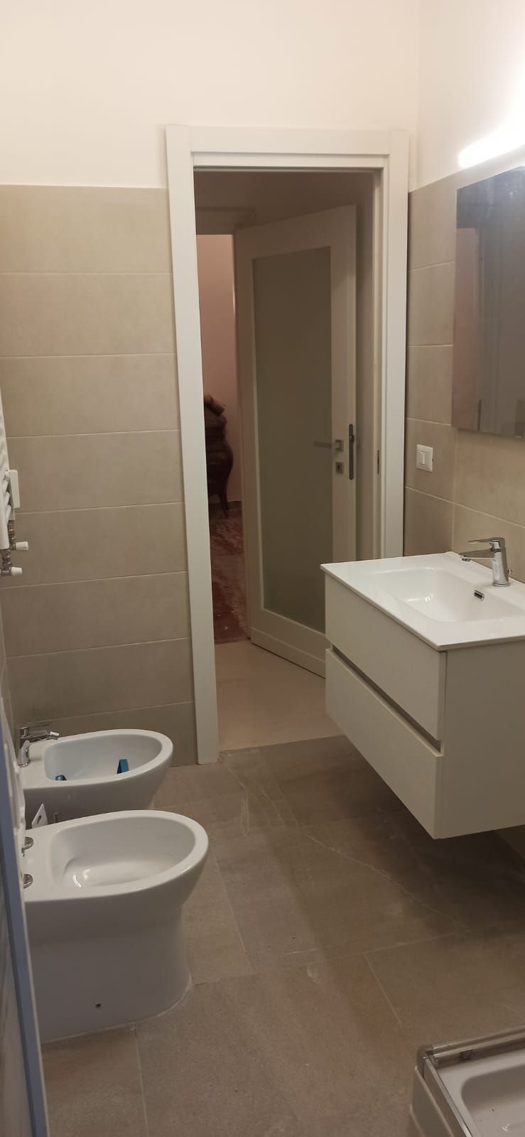 Bagno con WC, bidet, lavandino e porta aperta. Pareti e pavimento beige.