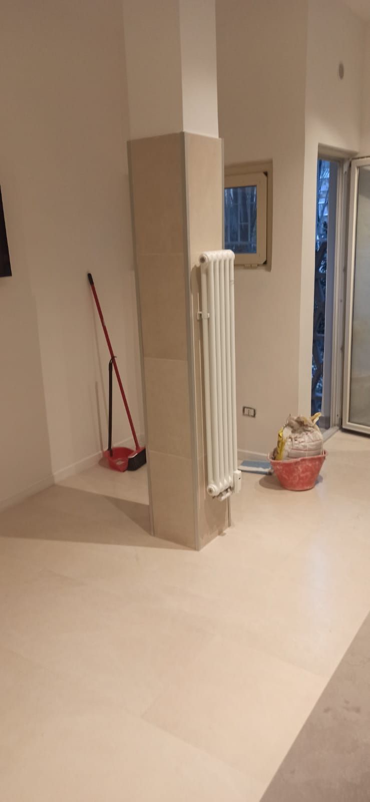 Ripresa dell'interno di una stanza con un pilastro centrale, un termosifone, una scopa e un cestino rosso. Pareti bianche e pavimento chiaro.