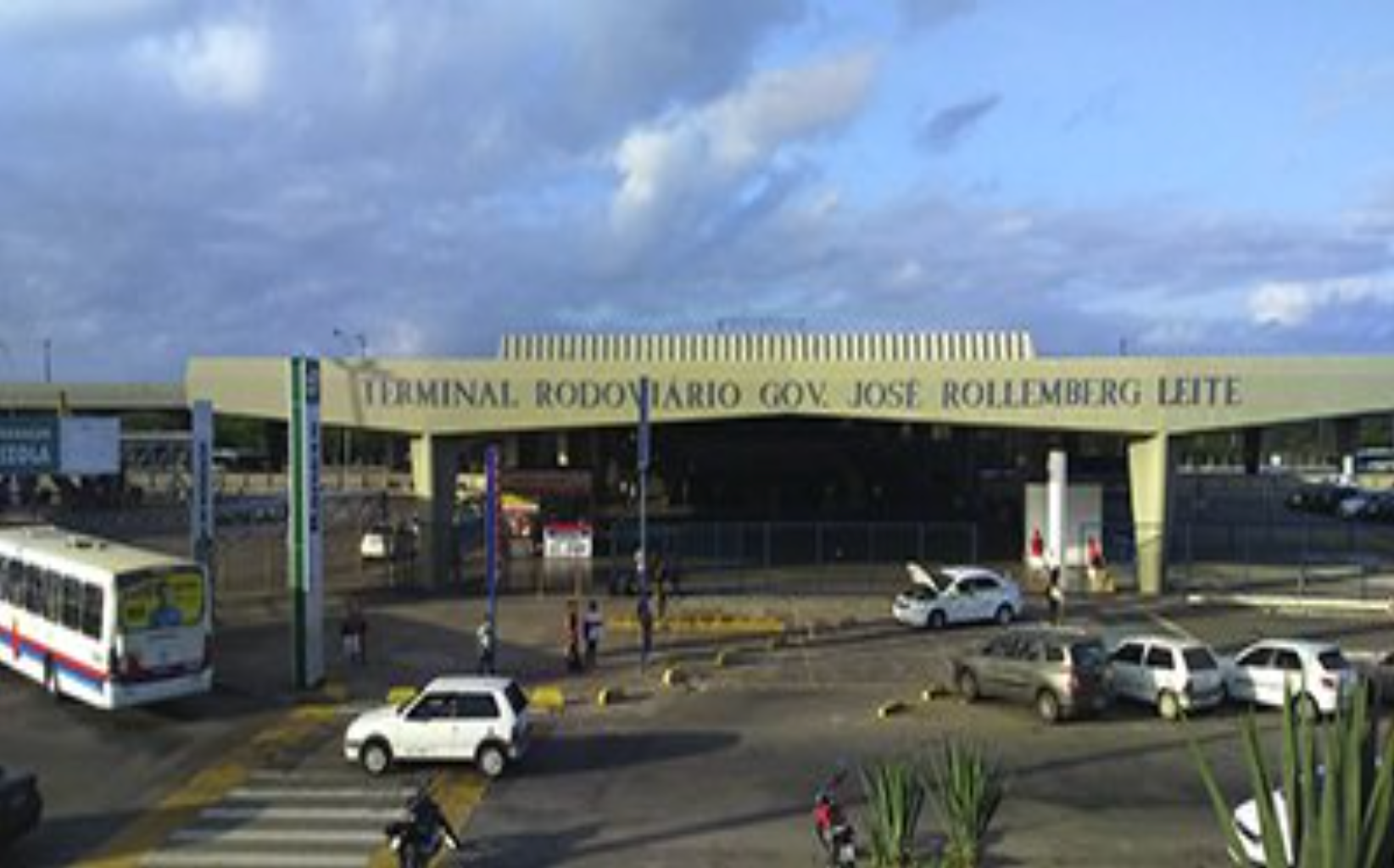 Entrance to Terminal Rodoviário Gov. José Rolemberg Leite with buses, cars, people, and a cloudy sky.