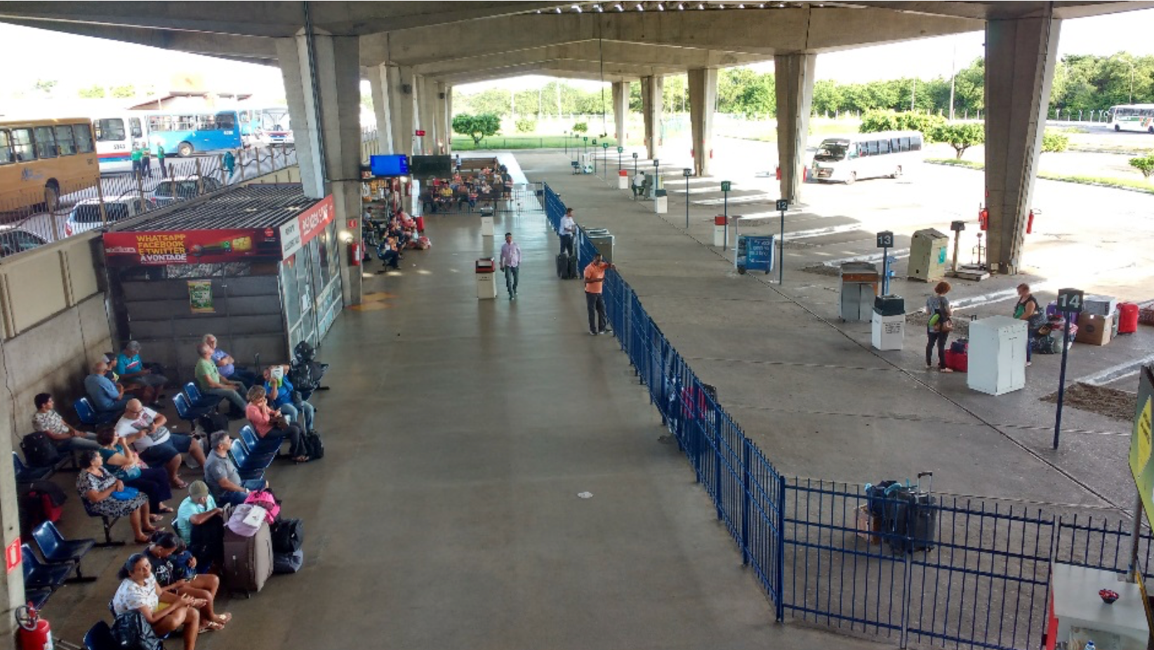 Terminal rodoviário: pessoas aguardando, área de espera com assentos, plataforma coberta, ônibus ao fundo.