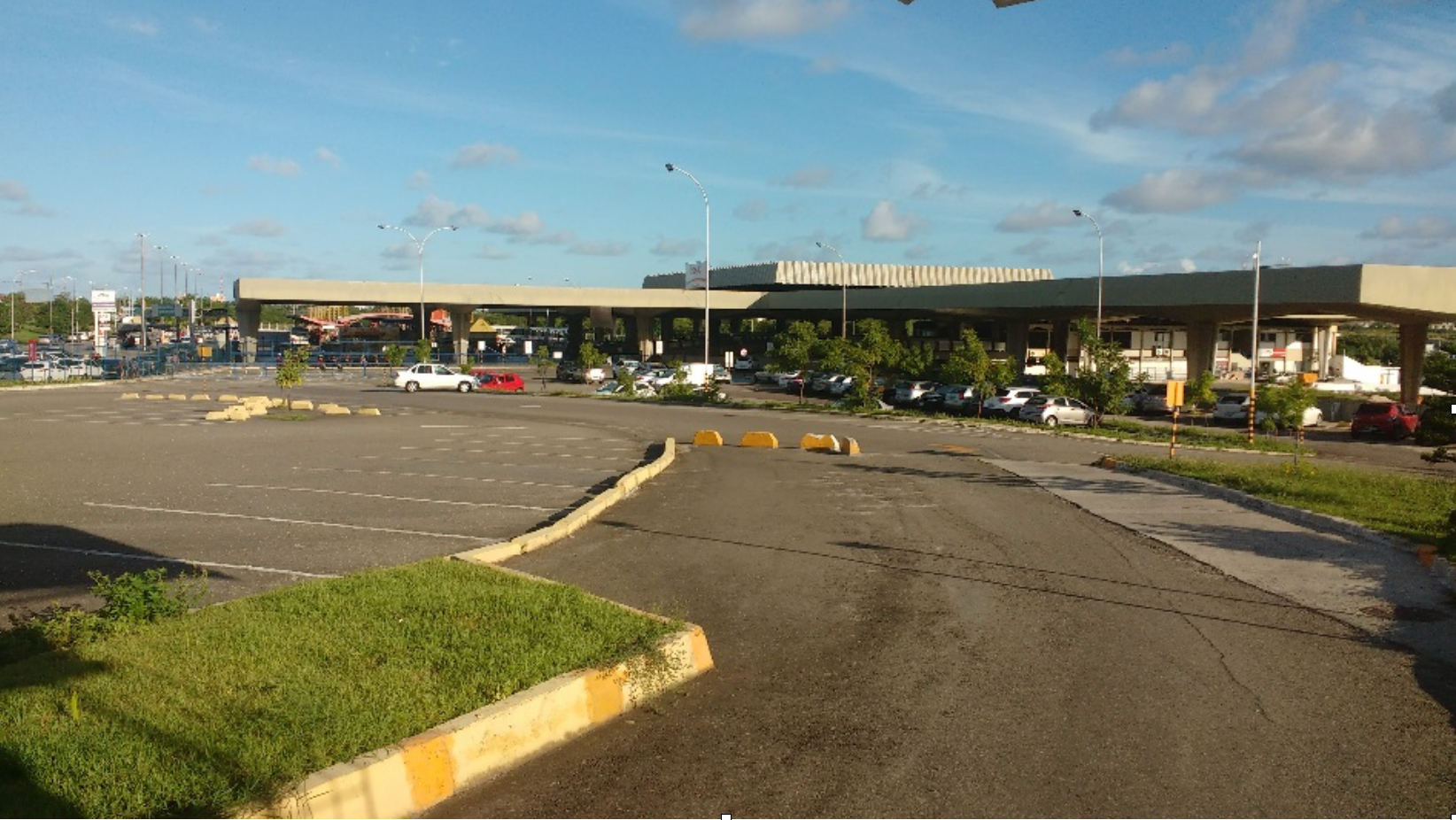 Uma praça de pedágio com carros enfileirados, dia ensolarado, céu azul e grama verde.