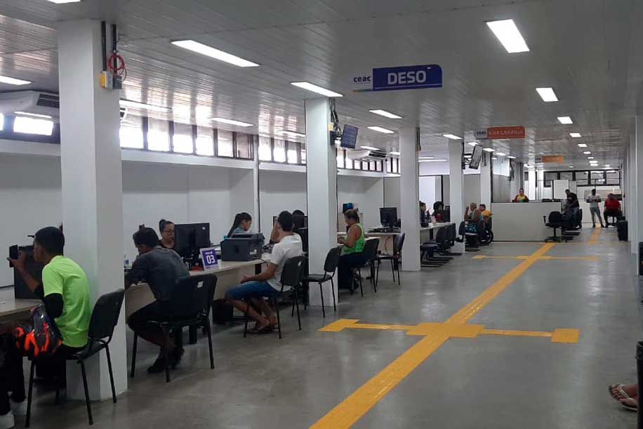 Pessoas em mesas de computador em um escritório bem iluminado. Linhas amarelas no chão, luzes no teto e placas informativas.