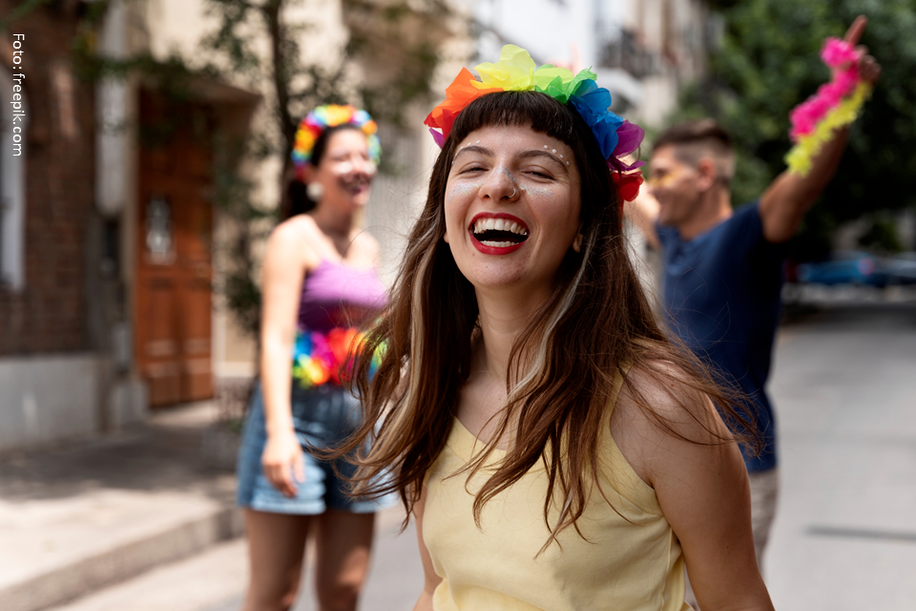 Mulher com coroa de arco-íris ri, outras pessoas ao fundo com roupas de arco-íris, ao ar livre.