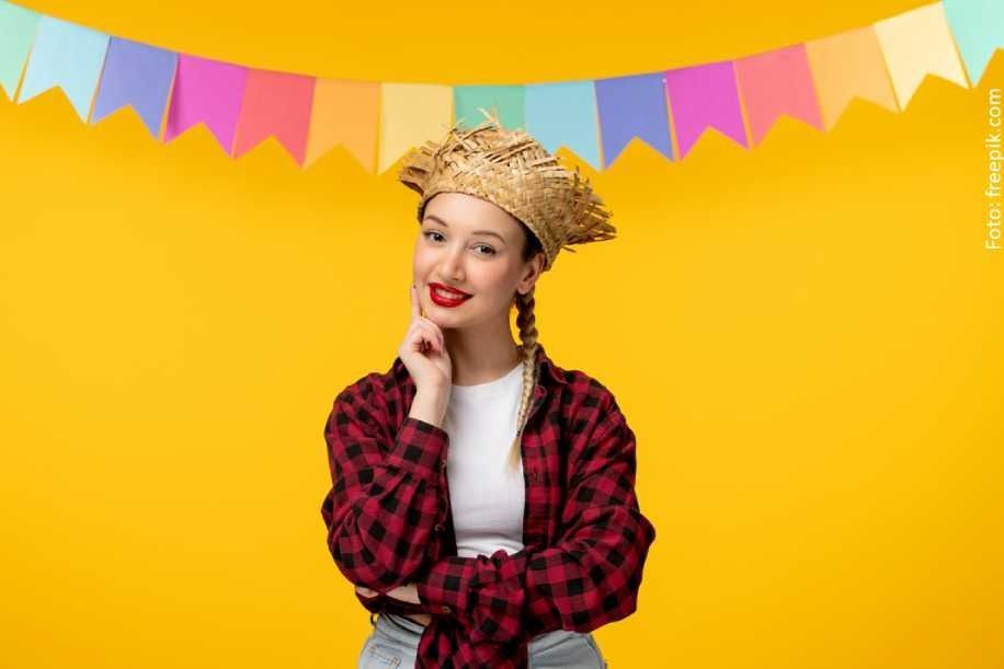 Uma mulher com chapéu de palha e camisa xadrez sorri contra um fundo amarelo com faixas coloridas.