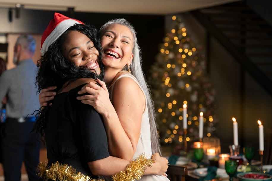 Duas mulheres se abraçando e sorrindo em uma festa de Natal; árvore e mesa iluminadas ao fundo.