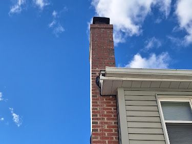 Brick chimney against a bright blue sky with scattered clouds, attached to a house with gray siding.