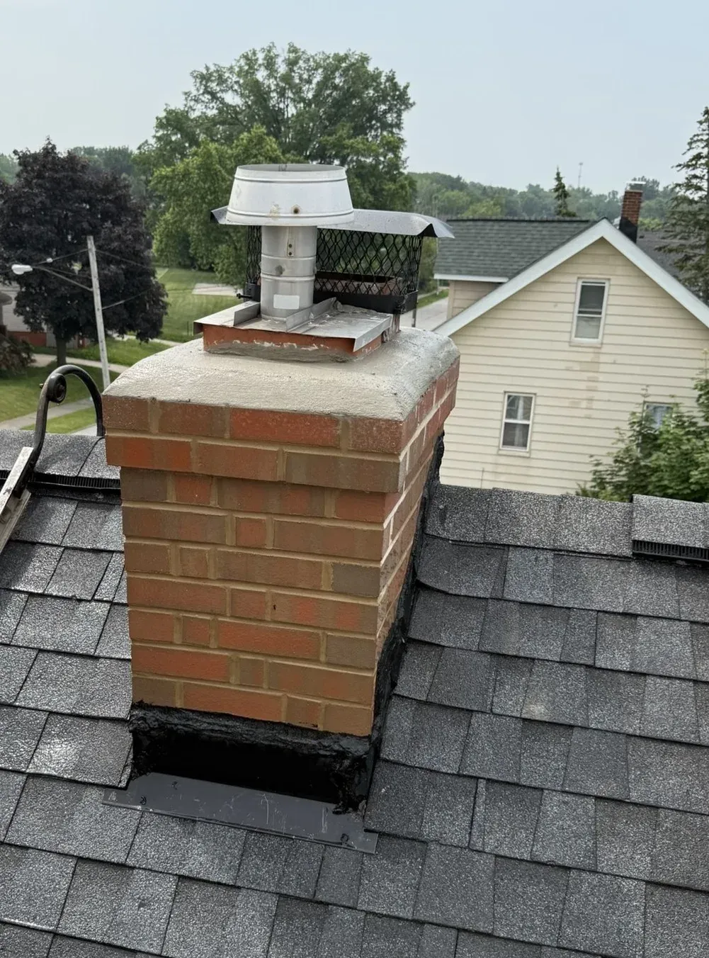 Brick chimney on a shingled roof, with a metal cap and flashing. The background shows trees and a house.