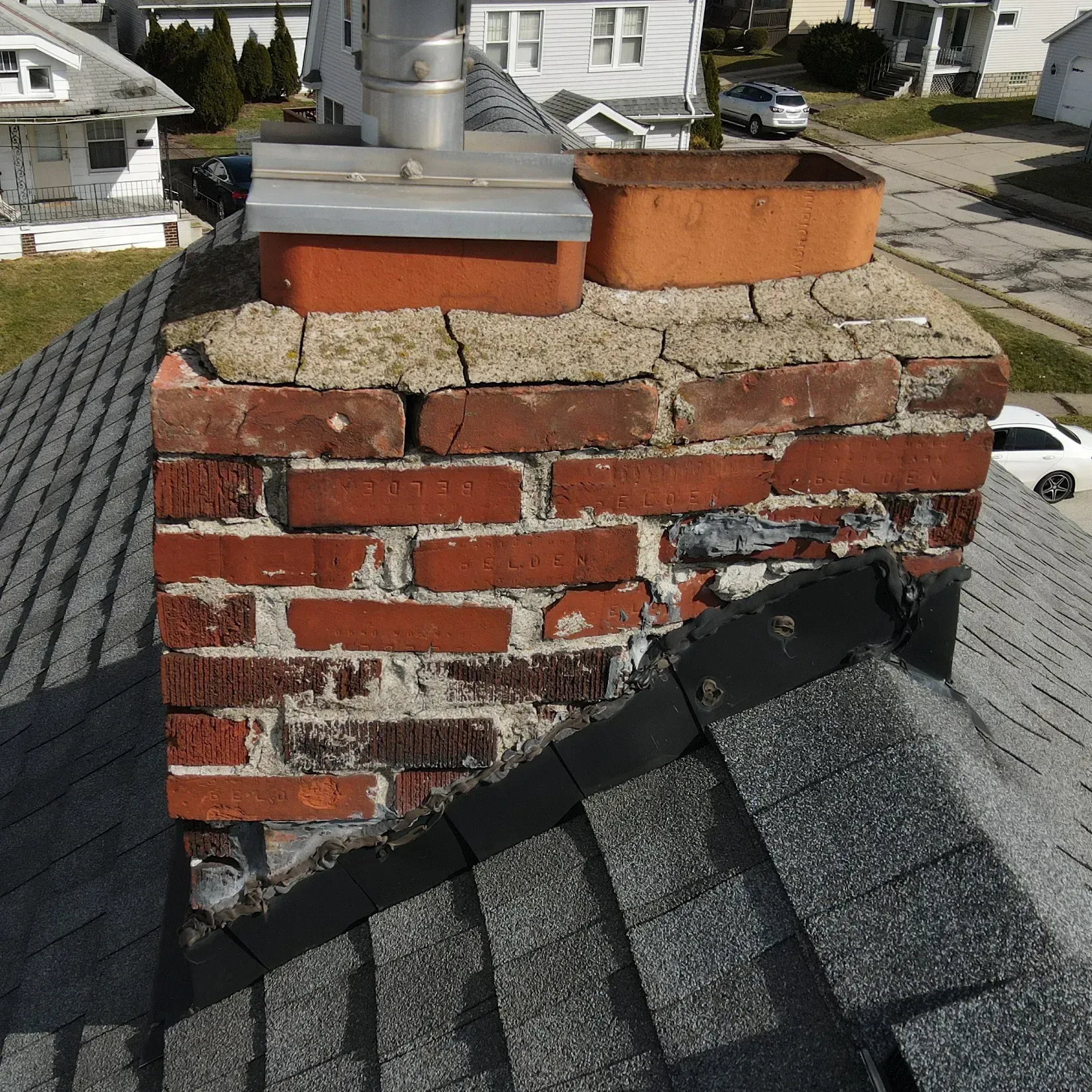 Brick chimney on a shingled roof shows cracked mortar and deterioration. The roof is in a residential area.