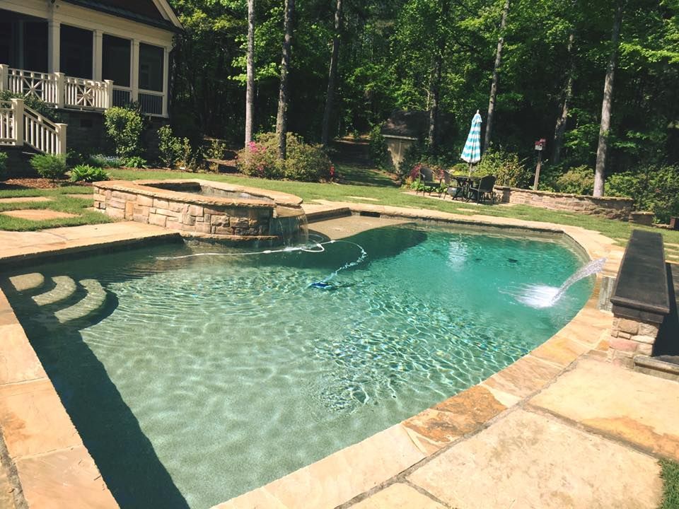 Pool with stone coping, waterfall feature, and a spa surrounded by a manicured lawn and trees.
