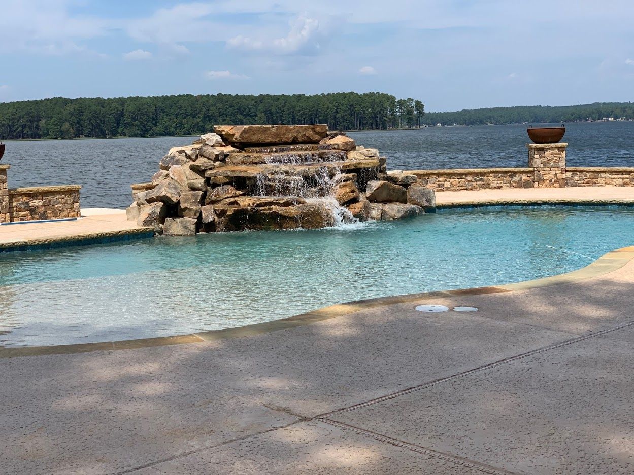 Pool with waterfall feature overlooking a lake. Blue water, stone details, and green trees in the background.