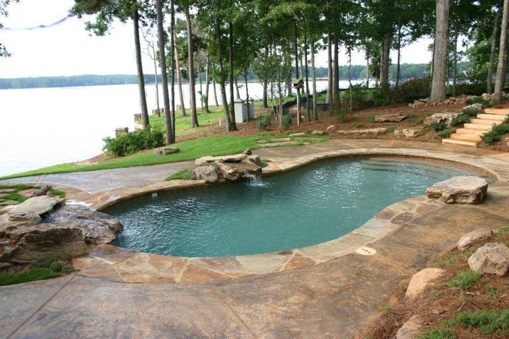 Pool with a waterfall by a lake, surrounded by stone patio and steps.