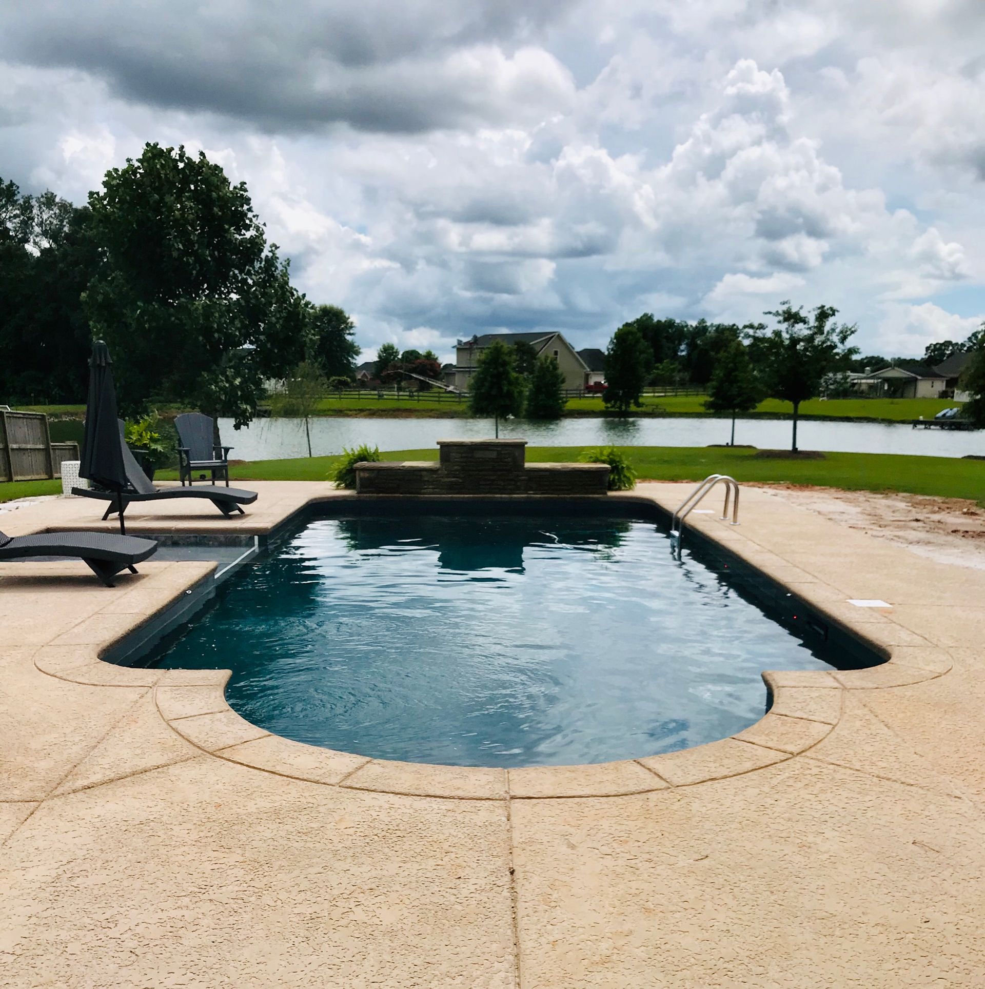 Swimming pool with dark water, lounge chairs, and view of a lake under cloudy skies.