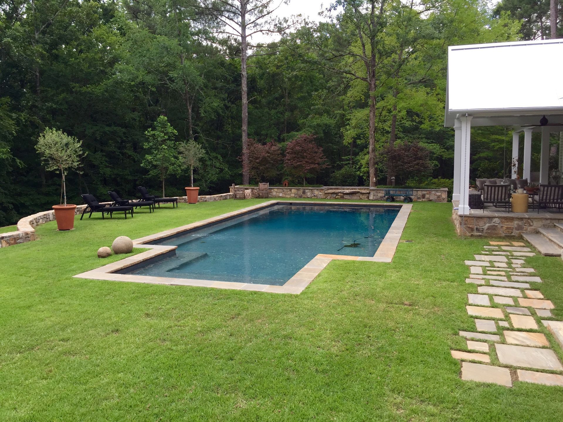 A rectangular pool surrounded by green grass and a stone pathway, with a white-roofed structure nearby.