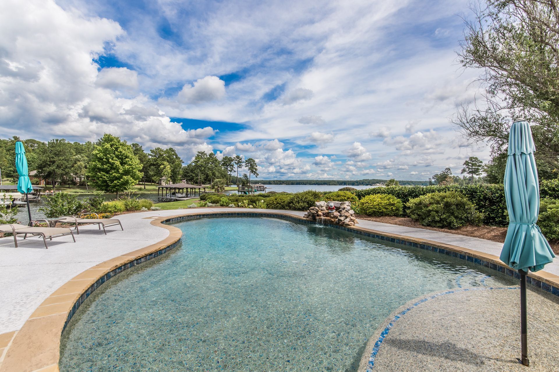 Pool with a view of a lake under a cloudy sky. Blue umbrellas and lounge chairs are nearby.