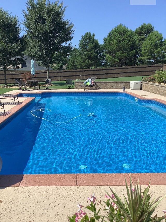 Swimming pool with blue water, surrounded by a concrete deck and a brown coping. Trees in the background.