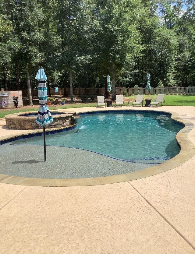 Pool with umbrella, fire pit, and patio surrounded by trees and grass.