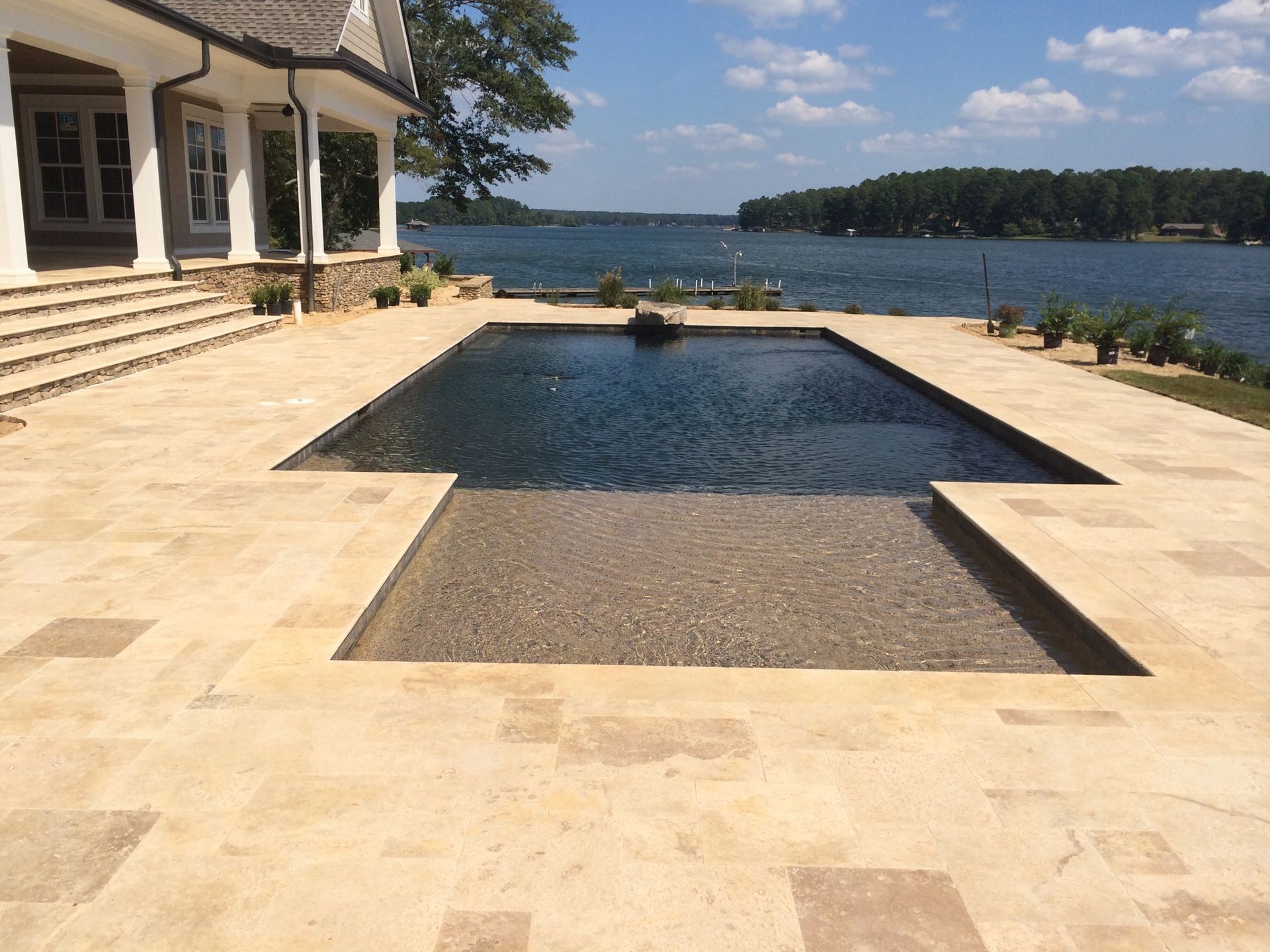 Pool overlooking a lake; beige stone patio, dark blue water. House in background.